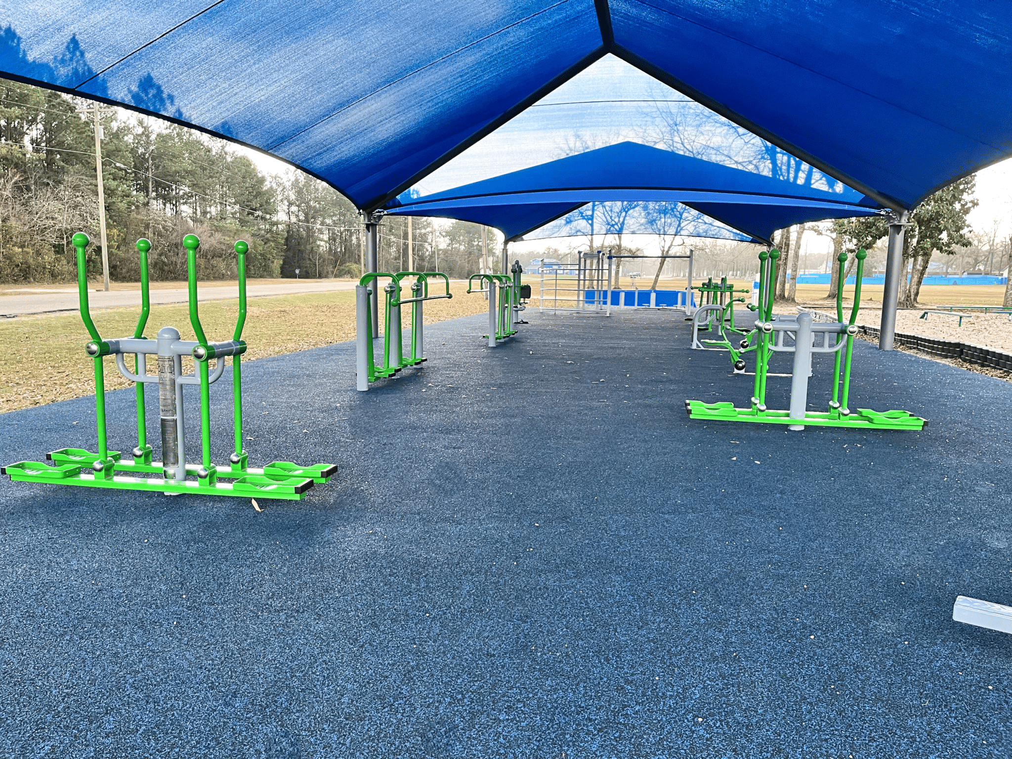 An outdoor physical education classroom. The ground is blue from poured-in-place rubber, and shows green ExoKids exercise equipment pieces. The area is covered with a blue shade structure.