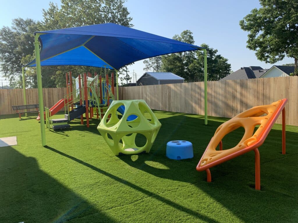 The image shows an outdoor early childhood playground covered with a blue shade structure. The playground equipment pieces are orange, lime green, and blue. The playground sits on top of turf grass.
