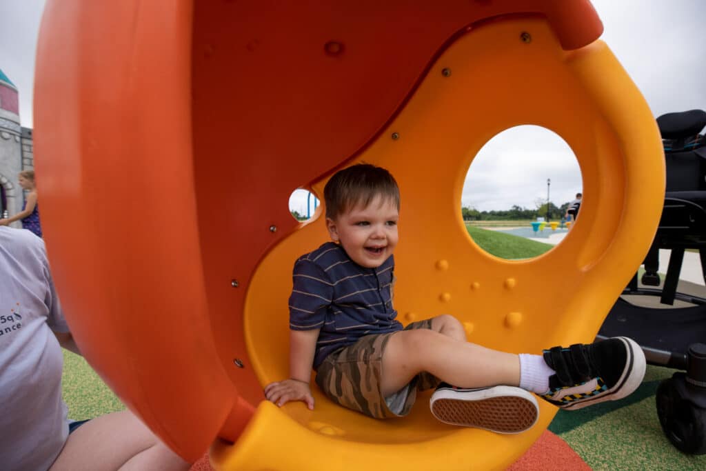 A young child sits in a Cozy Cocoon with his legs crossed. The Cozy Cocoon is orange and yellow. An adult sits to the child's right, out of frame.