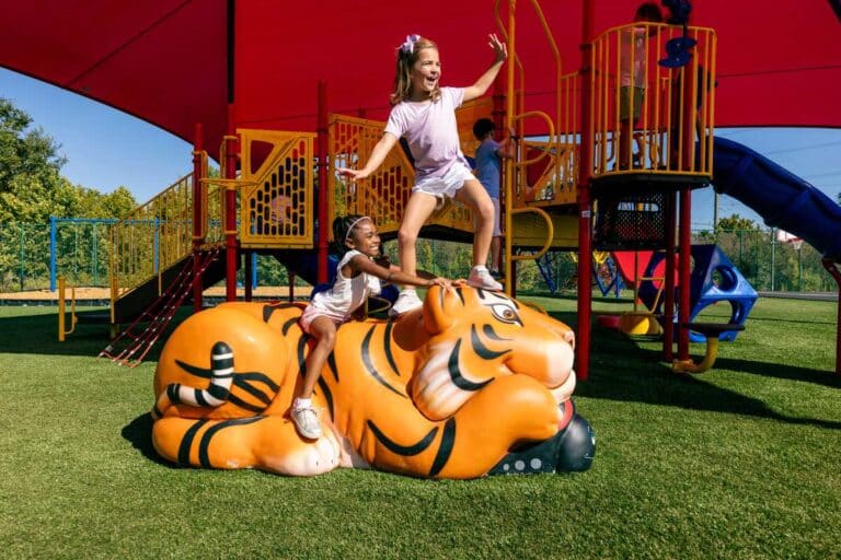 Two girls play on a sculptural tiger on a playground. One girl stands on the tiger's head.