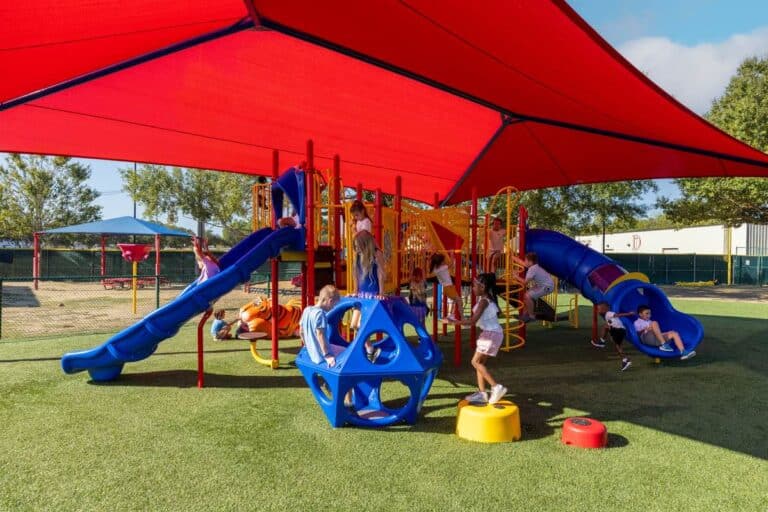 A large red shade structure hangs over a blue and yellow playground. Multiple children play on the playground.